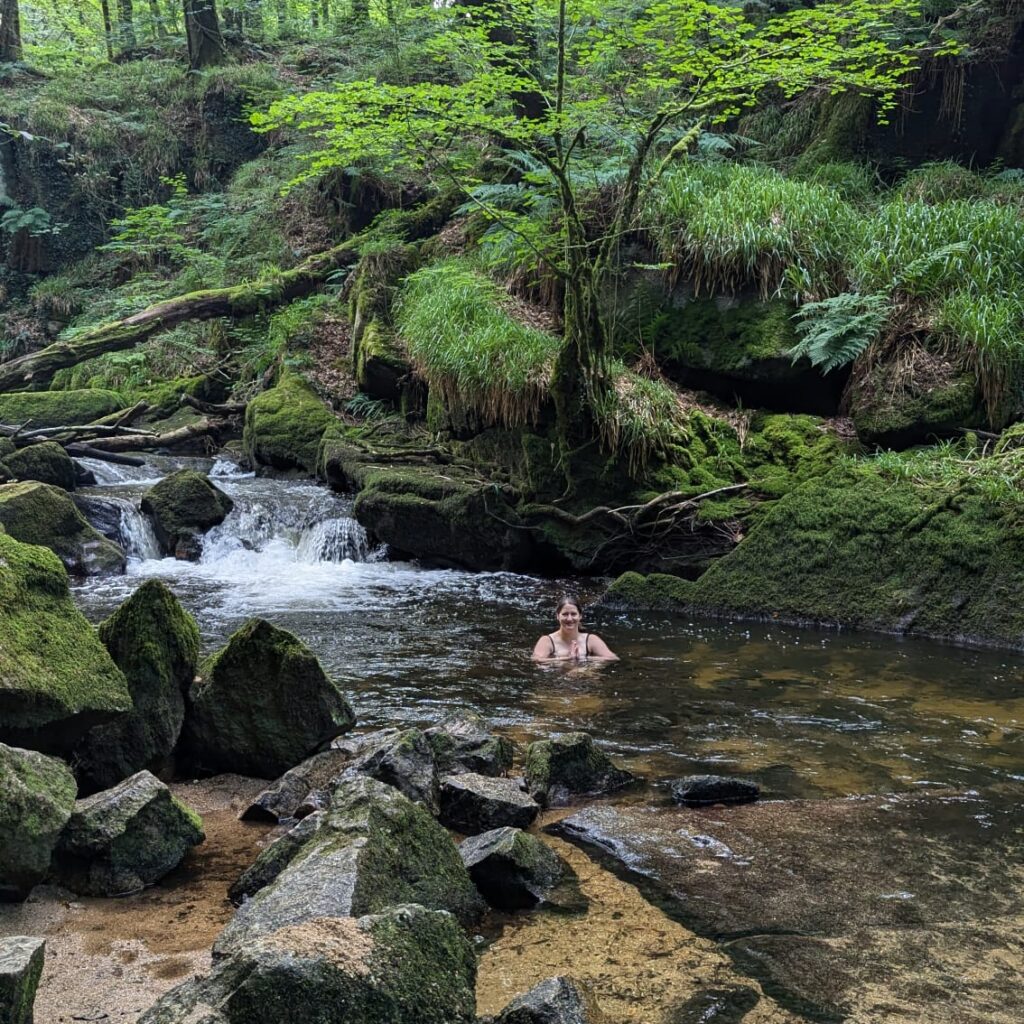 An image of the founder, Jo, dipping in a stream in front of a small waterfall surrounded by woodland.